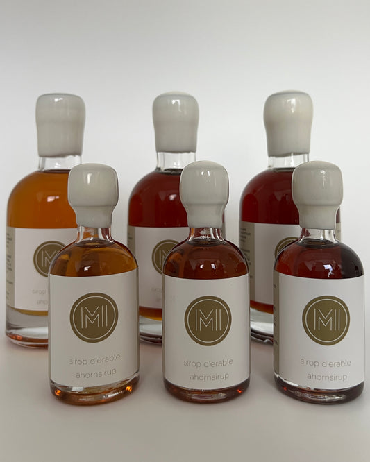 2 rows of side by side bottles of golden, amber and dark maple syrups, the first row are the smaller bottle size and the second row is the bigger bottle