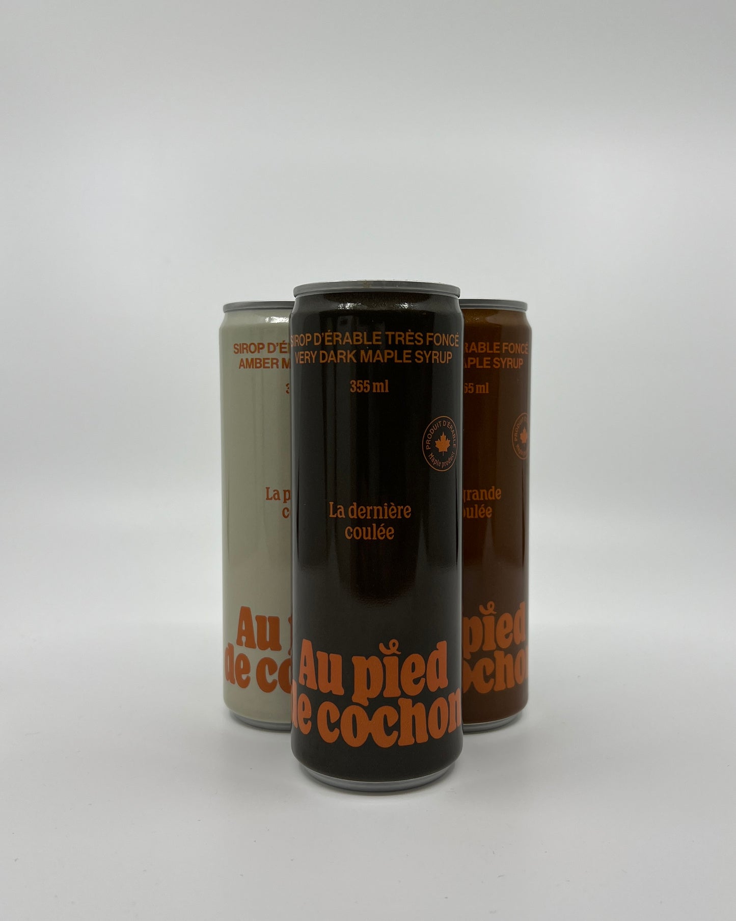 a bottle of very dark maple syrup 355ml named la dernière coulée without lid on a white background in front of 2 other bottles of the same style but different colors.