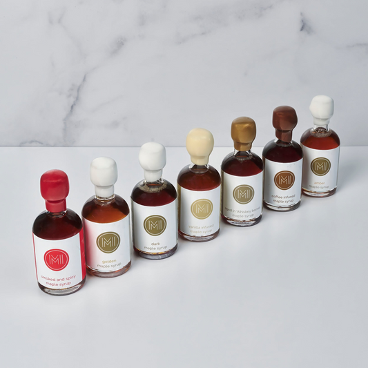 Six small glass bottles omaple syrups arranged in a row on a white surface against a light marble-patterned background.