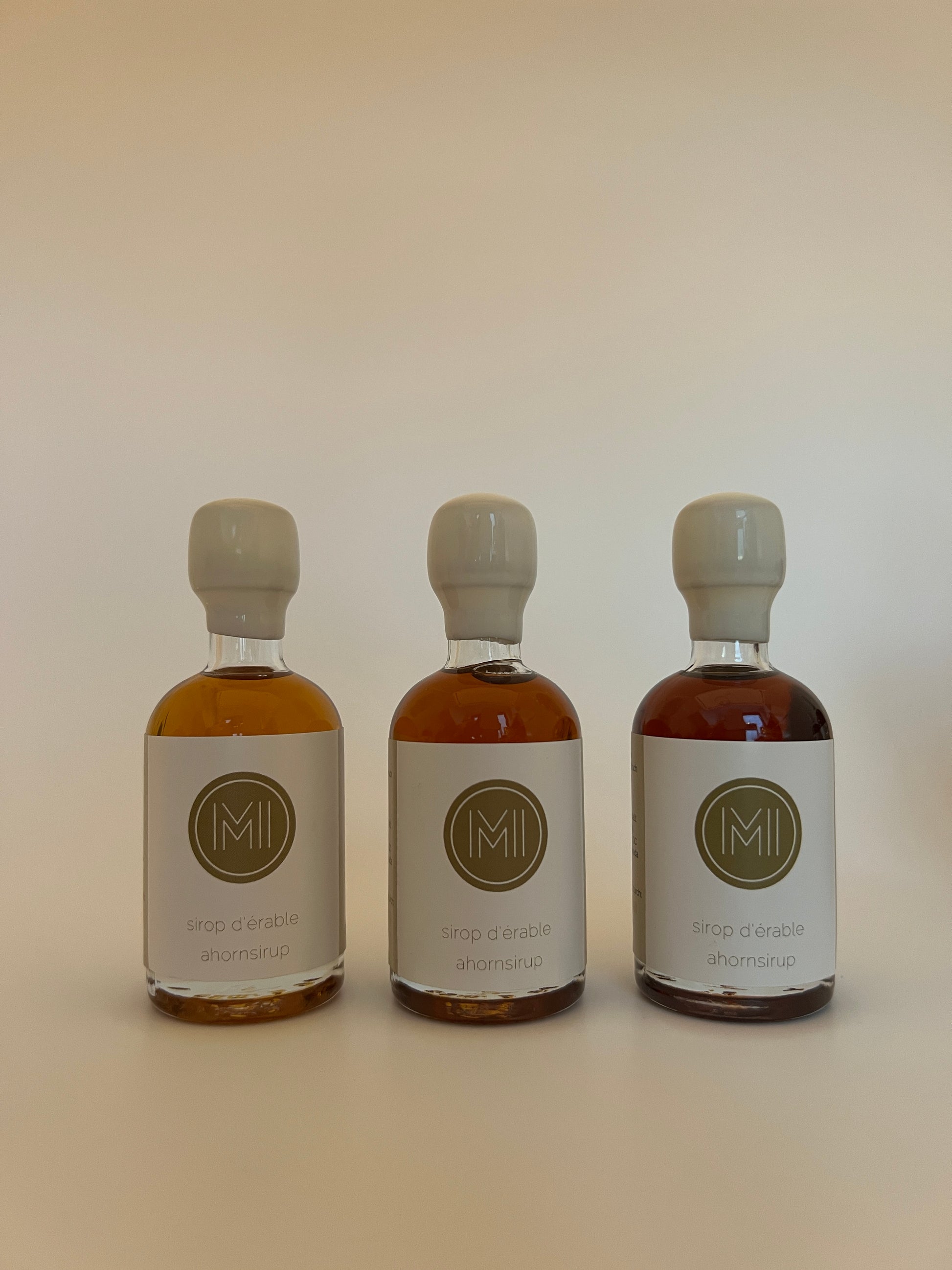 3 small bottles of golden, amber and dark maple syrup with white seals and logos all aligned on a white background