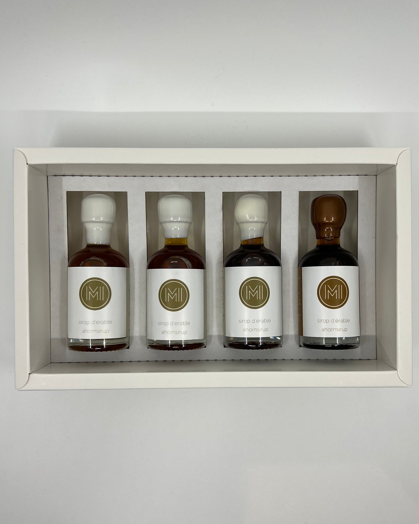 four side by side small bottles, three classic maple syrups and one coffee infused maple syrup with brown wax presented in a white box with insert on a white background