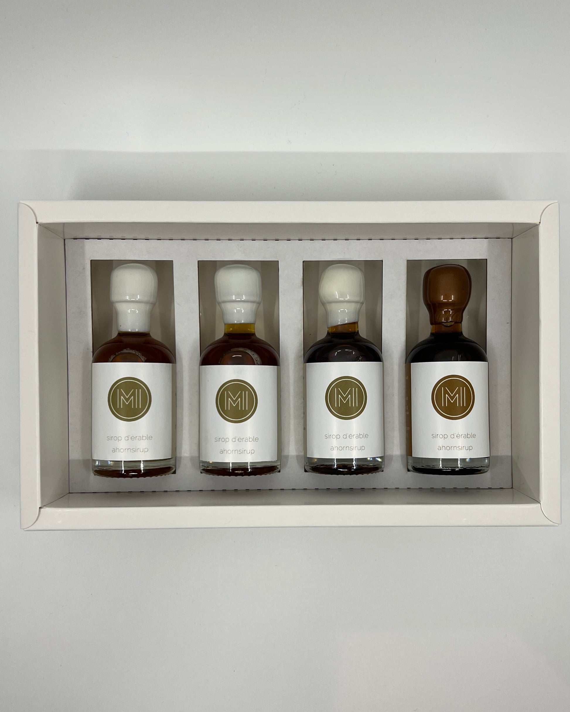 four side by side small bottles, three classic maple syrups and one coffee infused maple syrup with brown wax presented in a white box with insert on a white background