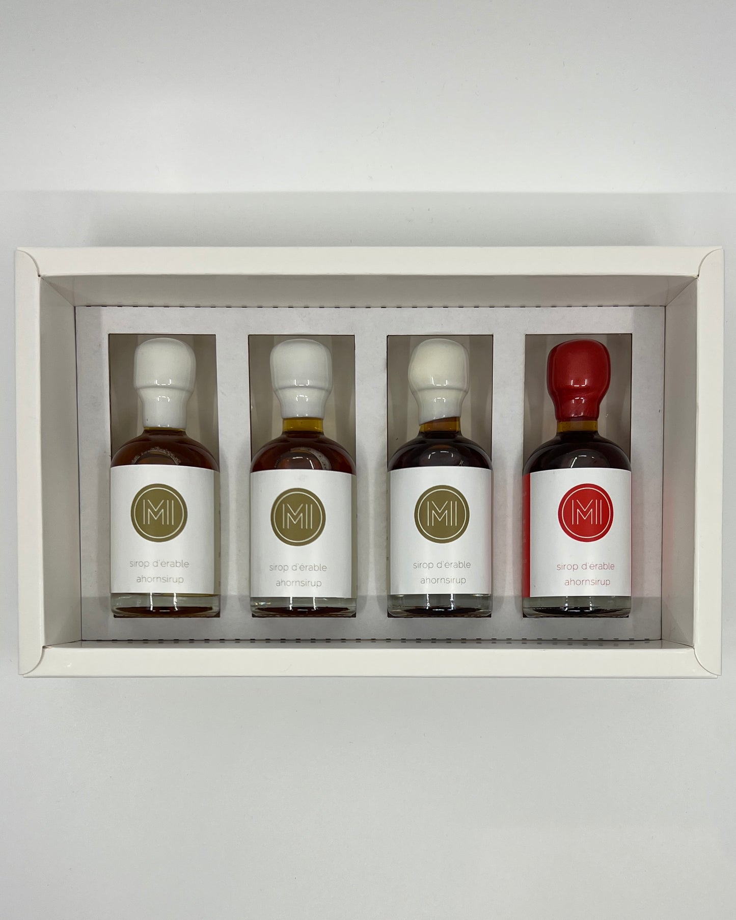 four side by side small bottles, three classic maple syrups and one smoked and spicy maple syrup with red wax presented in a white box with insert on a white background