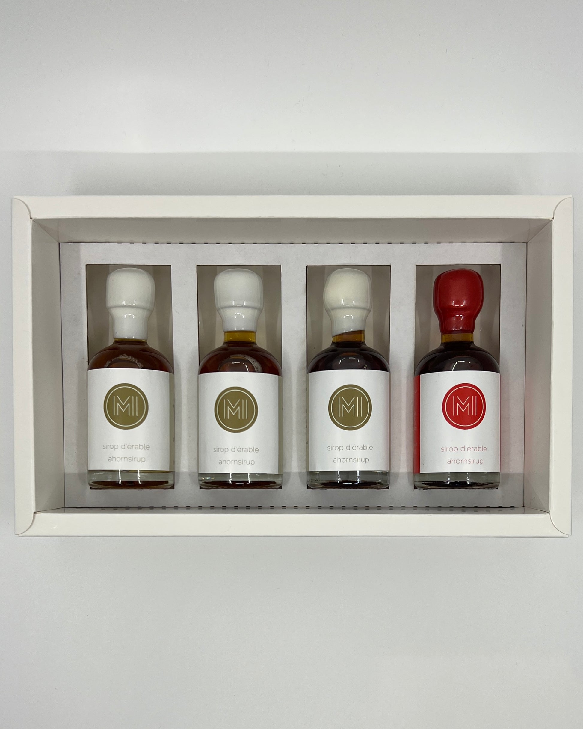 four side by side small bottles, three classic maple syrups and one smoked and spicy maple syrup with red wax presented in a white box with insert on a white background