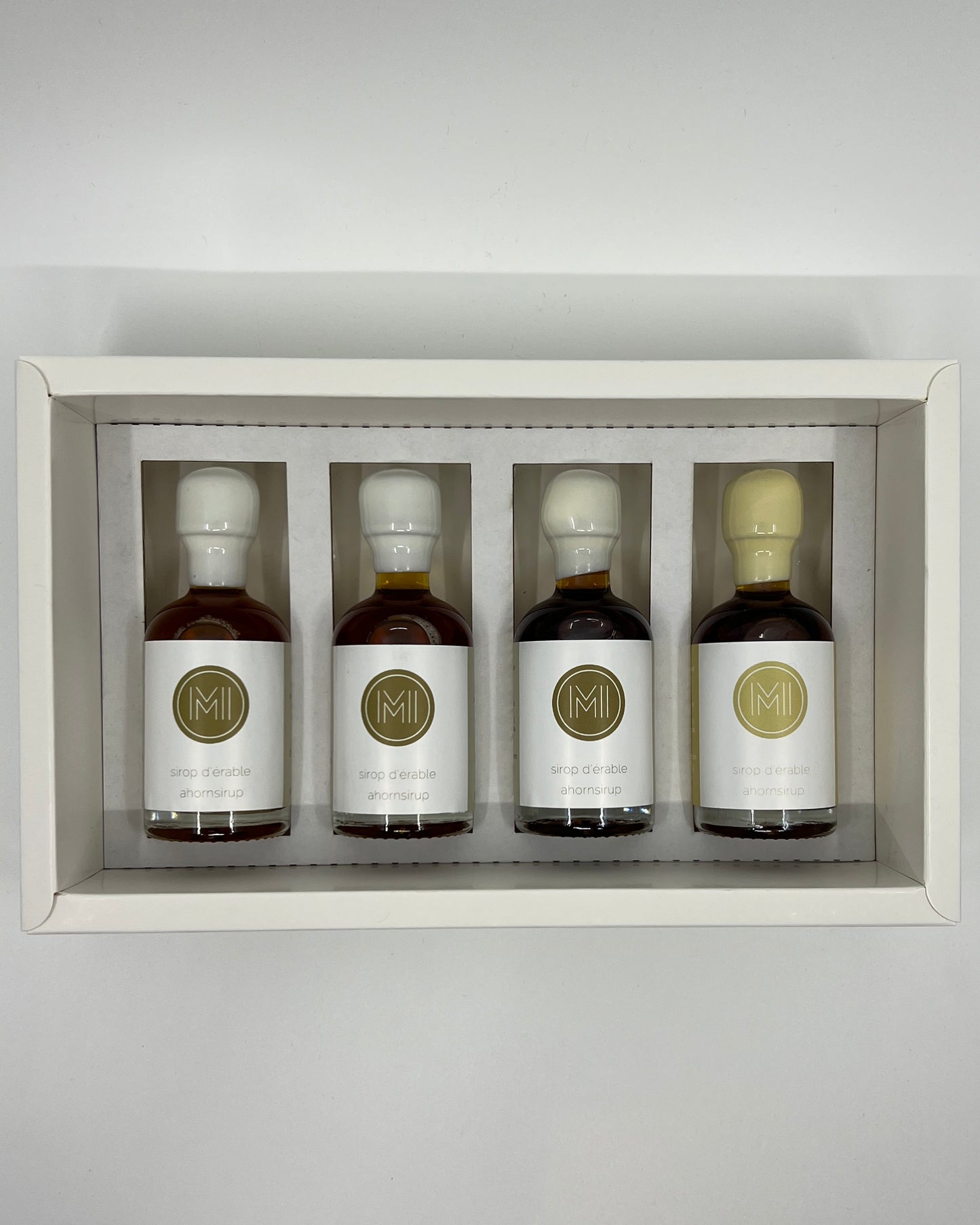 four side by side small bottles, three classic maple syrups and one vanilla infused maple syrup with beige wax presented in a white box with insert on a white background