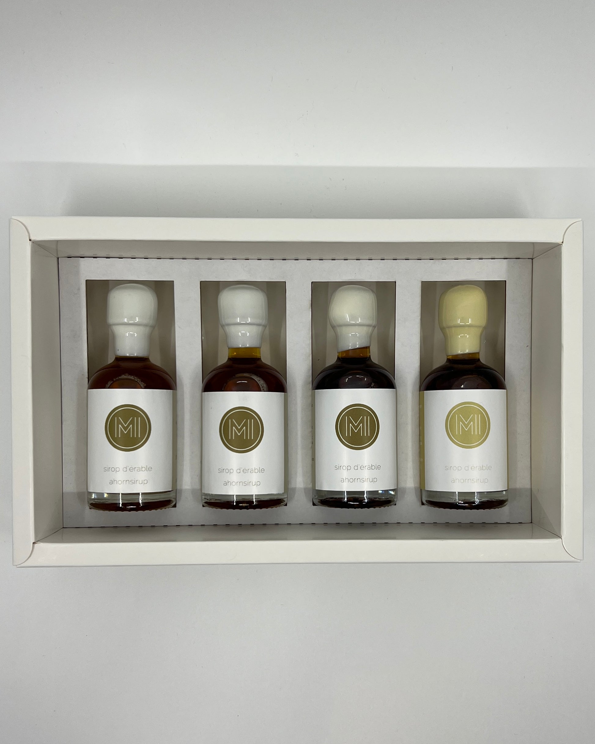 four side by side small bottles, three classic maple syrups and one vanilla infused maple syrup with beige wax presented in a white box with insert on a white background