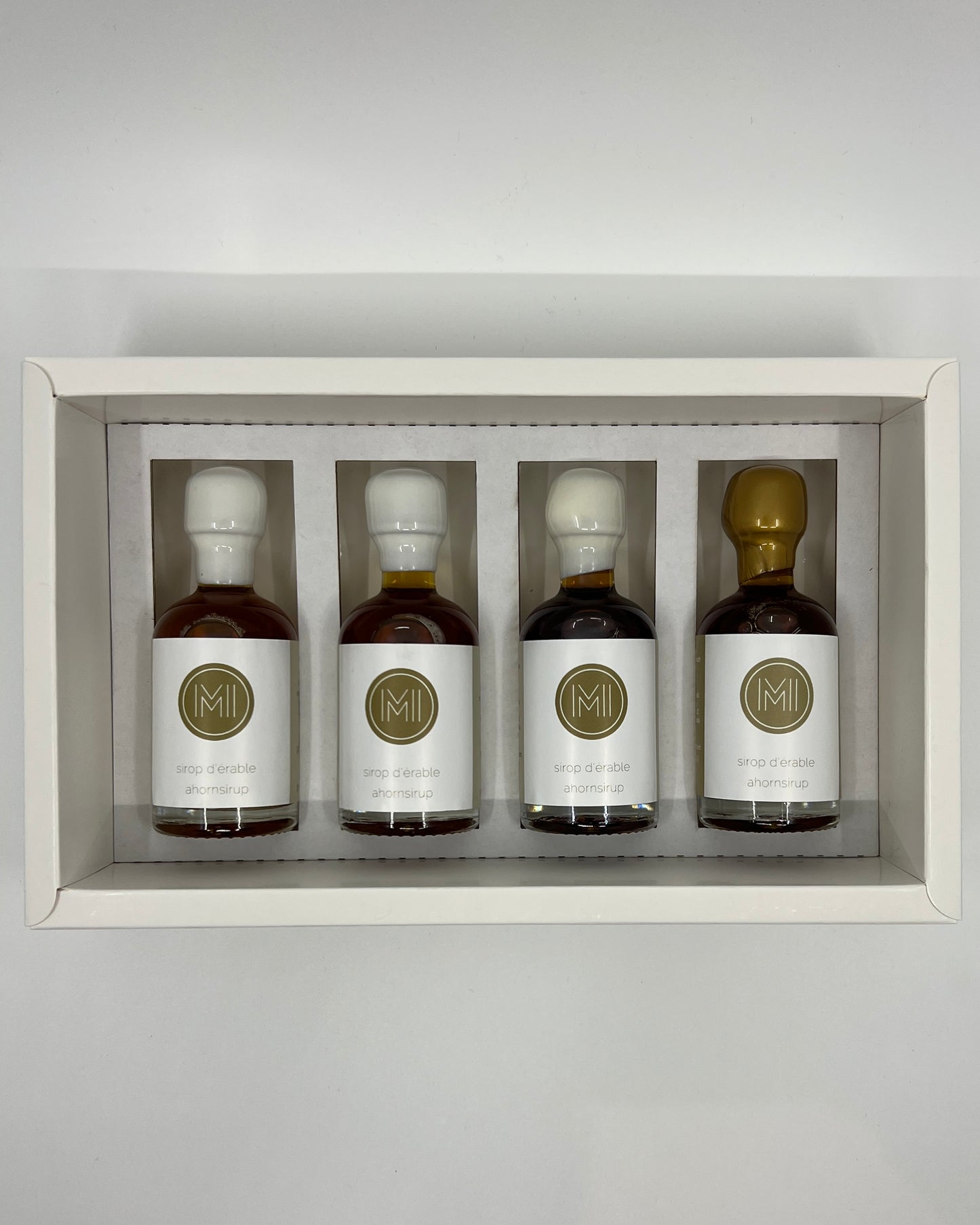 four side by side small bottles, three classic maple syrups and one aged in whiskey barrel maple syrup with gold wax presented in a white box with insert on a white background