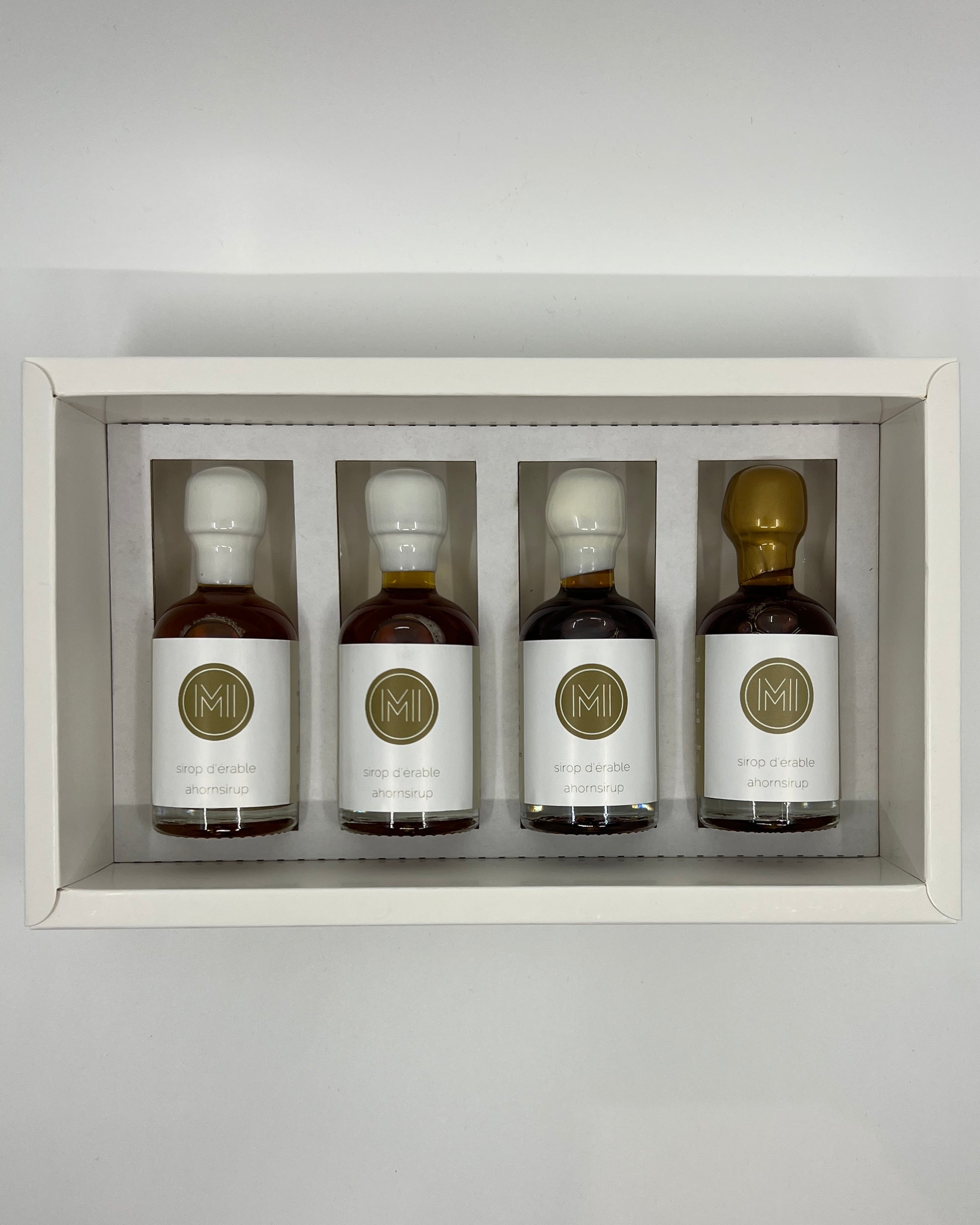 four side by side small bottles, three classic maple syrups and one aged in whiskey barrel maple syrup with gold wax presented in a white box with insert on a white background
