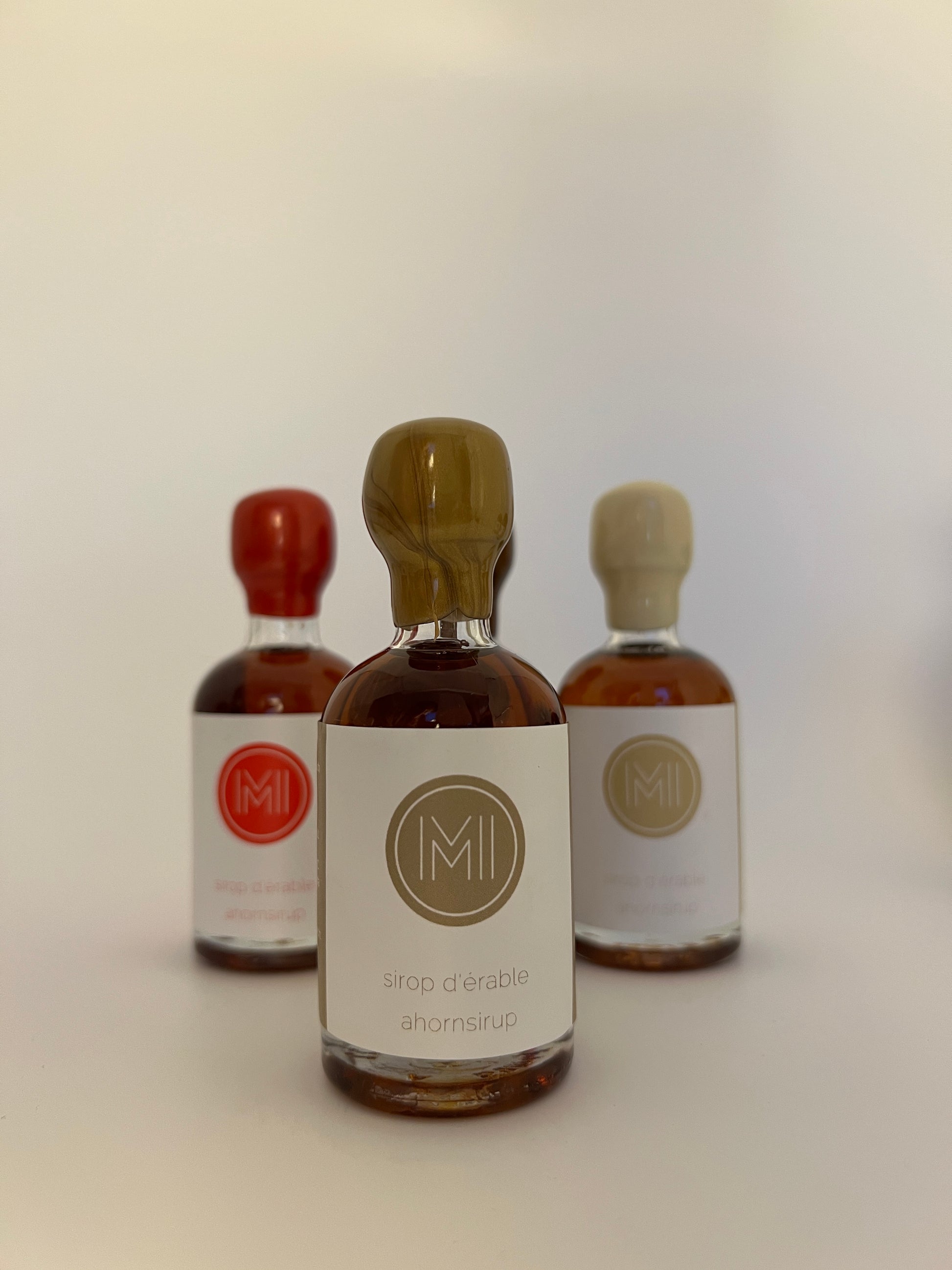 a 50 ml of aged in whiskey barrel maple syrup in bottle with a gold wax seal in front of 2 other bottles of the same style but different colors on a white 
