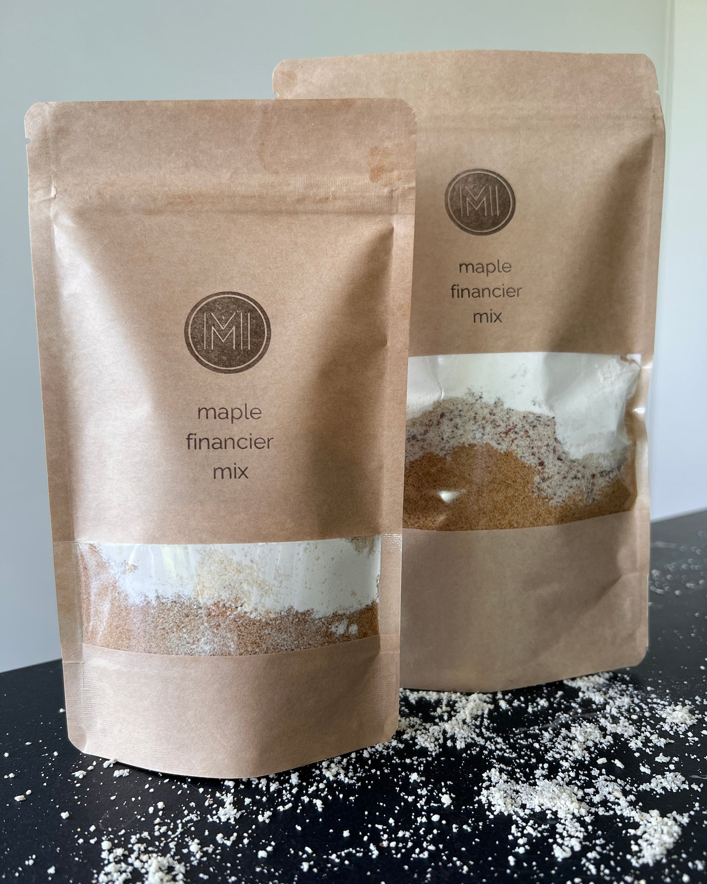 Two paper bags side by side on a countertop with see-through window showing maple financier mix made of maple sugar, almonds powder and flour