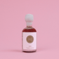 A small glass bottle of dark maple syrup with a white label and white cap, filled with dark reddish-brown syrup, centered on a solid pink background.