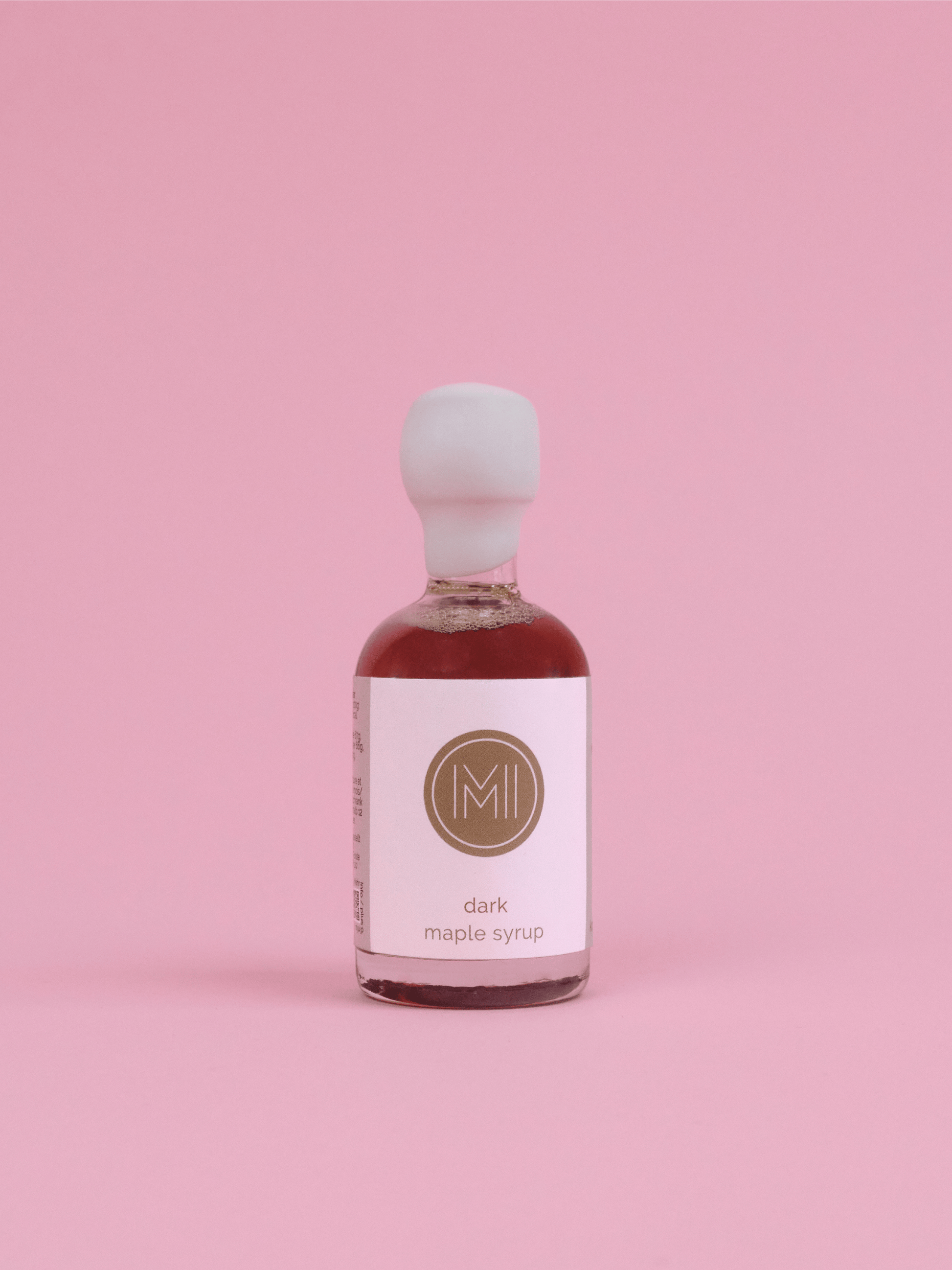 A small glass bottle of dark maple syrup with a white label and white cap, filled with dark reddish-brown syrup, centered on a solid pink background.