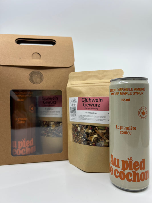 a bottle of 355 ml of amber maple syrup with in second plan a bag of mulled wine spices and in third plan a gif set brown cardboard box with see through window showing a bottle of dark maple syrup and another bag of mulled wine spices.