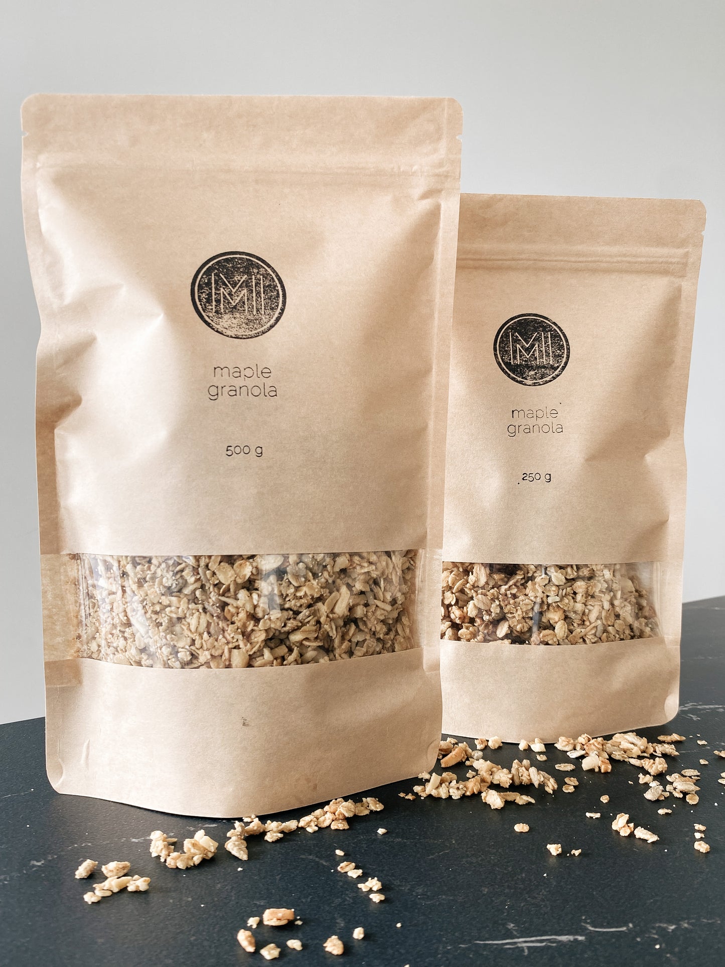 Two sizes paper bags (250g and 500g) with see-through window showing maple granola and granola sample on a grey countertop